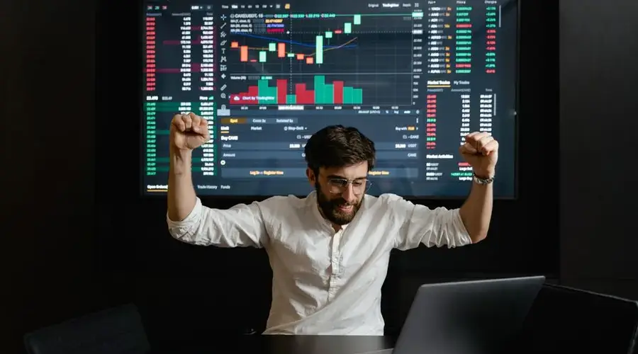 Businessman celebrates stock market success with hands raised in excitement at a trading desk.
