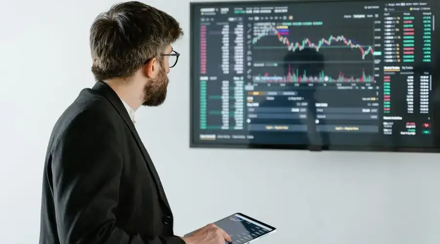Man observing stock market data on a screen and holding a tablet, representing financial analysis and trading.