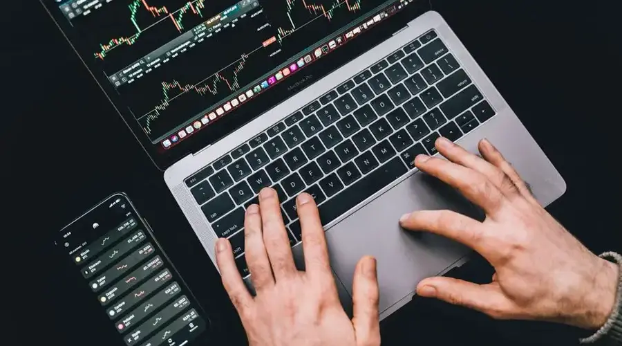 Hands typing on a laptop displaying stock market charts and a smartphone with financial apps.