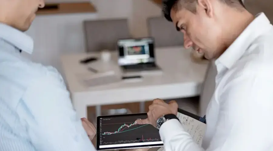 Two businessmen discuss stock market trends using a tablet with visible graphs.