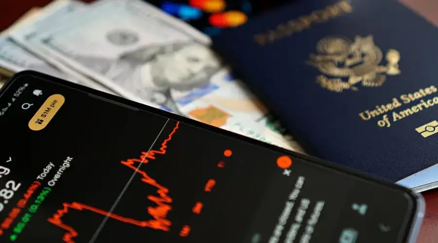 Close-up of smartphone with stock chart, cash, and US passport, symbolizing travel and finance.