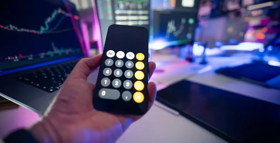 Smartphone with calculator app against financial charts on monitors, symbolizing financial planning.