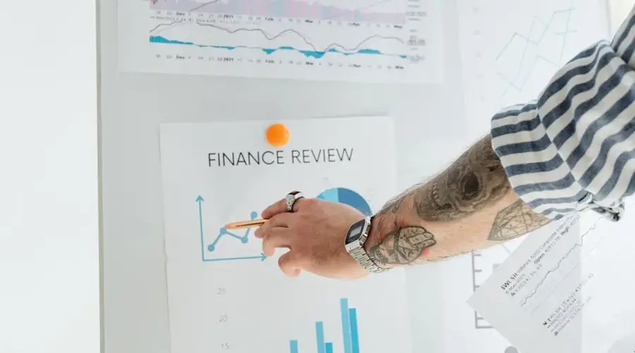 A person with tattooed arms presenting financial data using charts and graphs on a whiteboard.