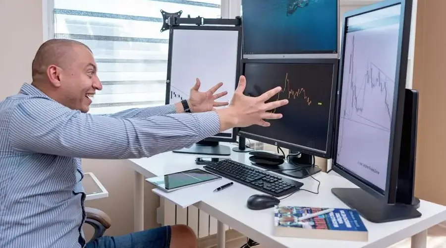 A joyful day trader celebrates a market victory in a modern office setup with multiple monitors displaying stock charts.
