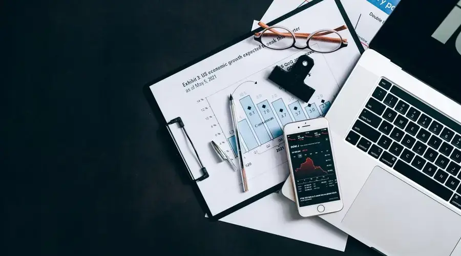 High angle view of an office desk with laptop, clipboard, and smartphone displaying stock market data.