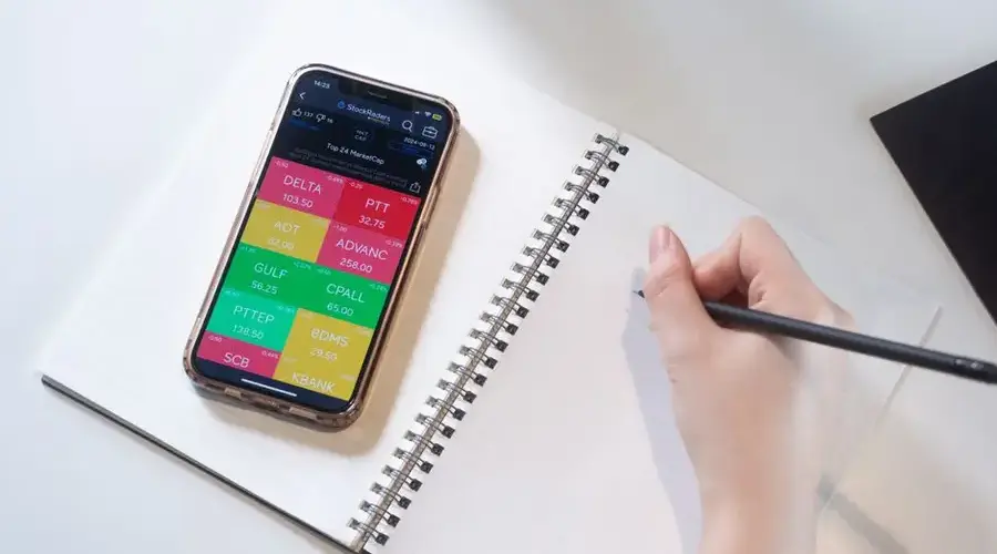 Close-up of a smartphone displaying stock data beside a hand writing in a notebook.