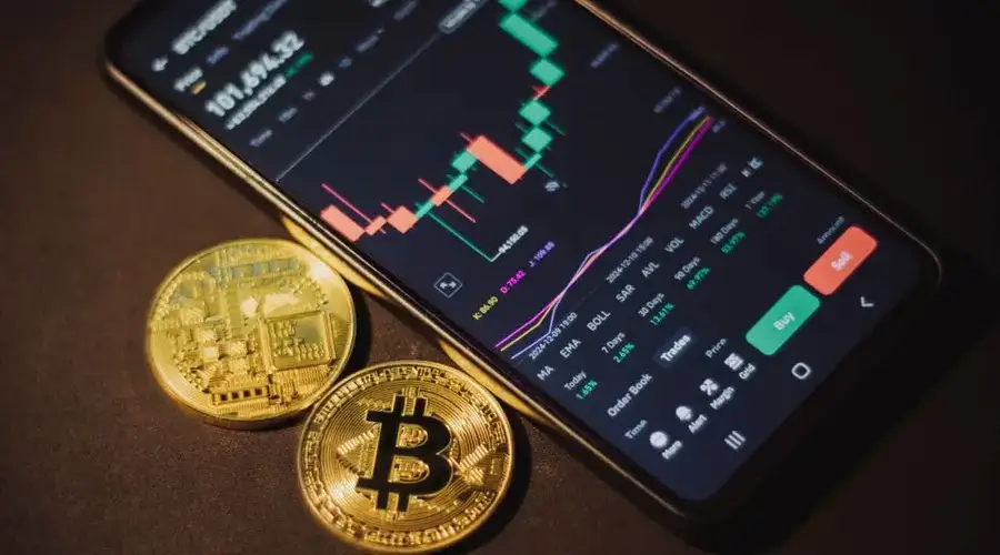 Smartphone displaying trading app with Bitcoin coins on a dark surface.