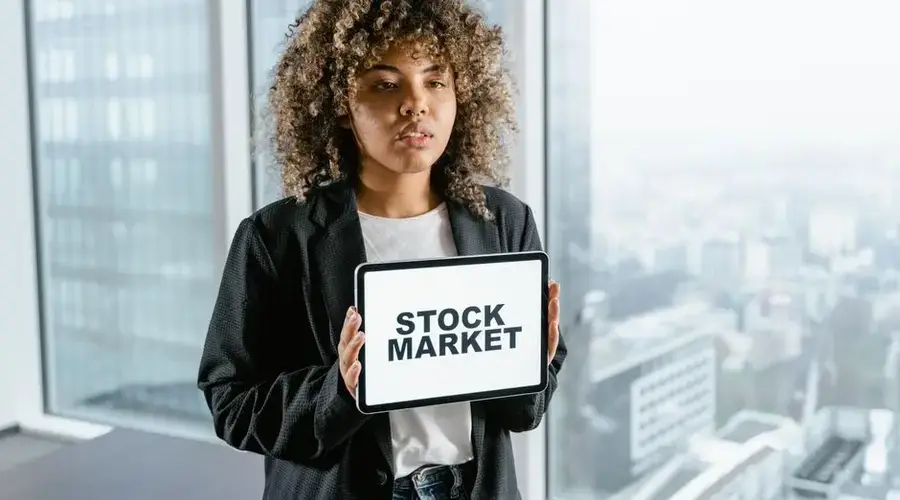 Woman in office holding a tablet with 'Stock Market' displaying on the screen, cityscape background.