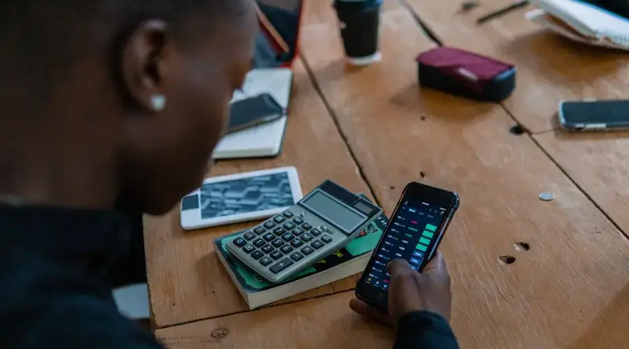 Close-up of individual using smartphone for financial data analysis, calculator nearby on wooden table.