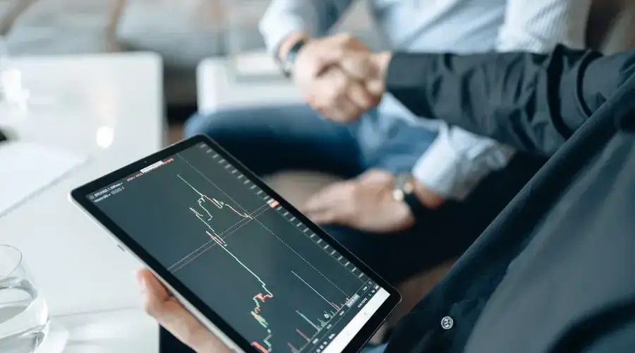 Two adults shaking hands with a stock market chart on a tablet, symbolizing a business deal.
