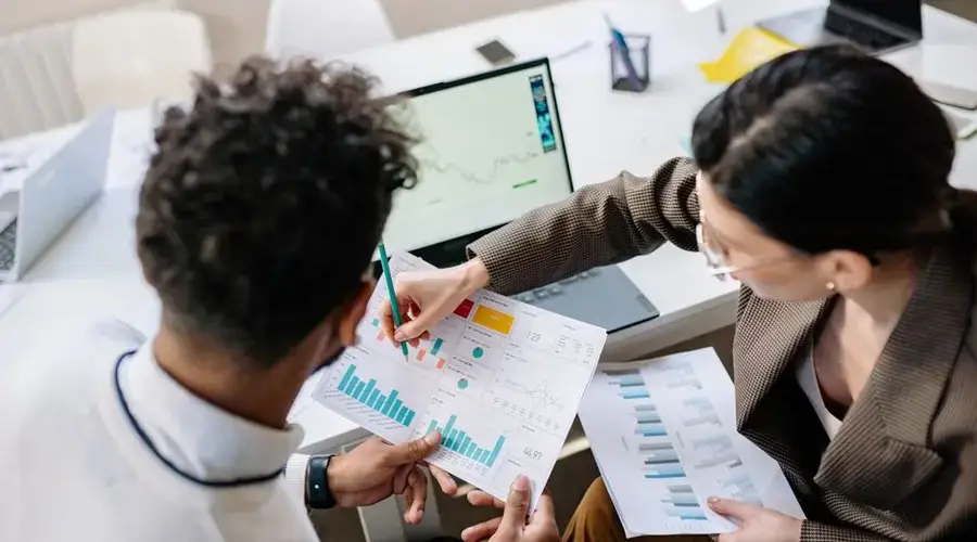 Two colleagues in a meeting room discussing financial charts and graphs on a laptop and paper.