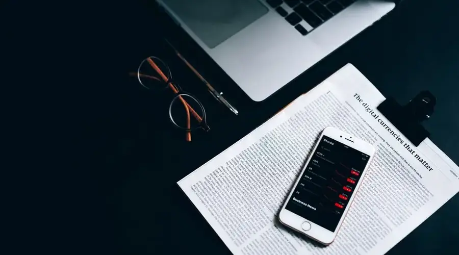 Flat lay of a digital workspace featuring a smartphone, glasses, and a laptop on a dark desk.