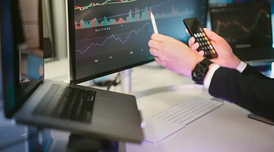 A professional analyzing financial charts on computer monitors and a smartphone, symbolizing digital finance strategies.