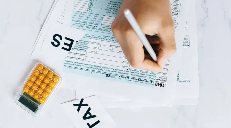 Close-up of hand writing on tax form with calculator nearby on white surface.