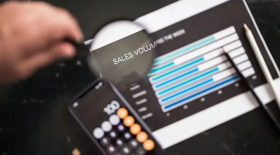 Close-up of a magnifying glass focusing on a sales volume chart next to a smartphone calculator app.