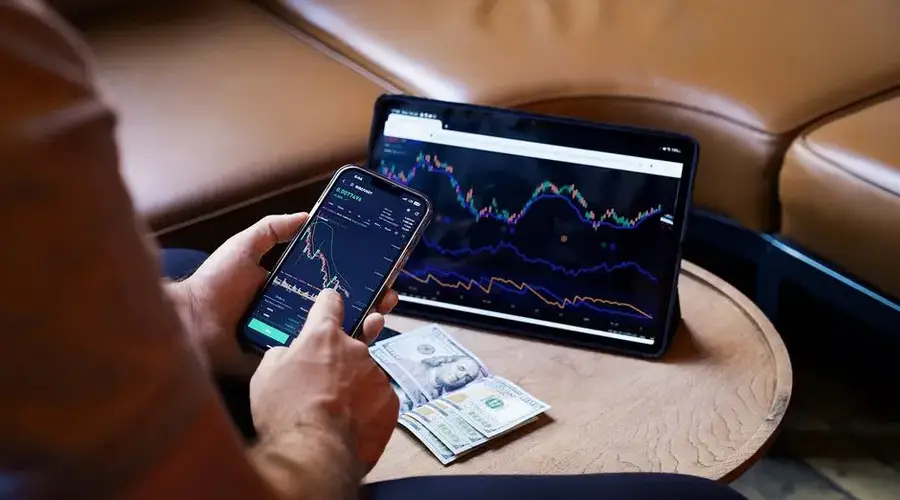 A person using a smartphone and tablet for stock market analysis, with graphs and cash visible.