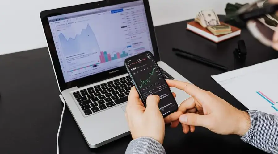 Person analyzing stock market data on a laptop and smartphone indoors.