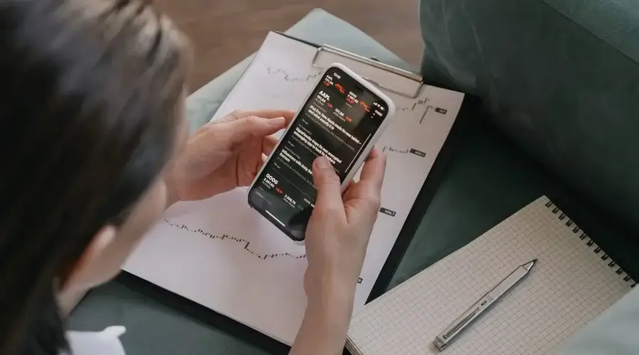A woman reviews financial data on her smartphone with charts and graphs around her for stock market analysis.