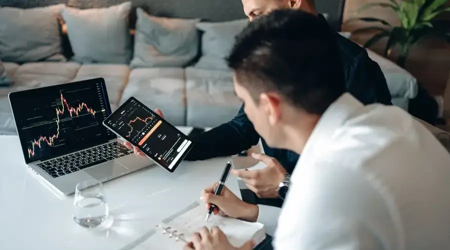 Two men discussing market trends using a tablet and laptop in a modern office.