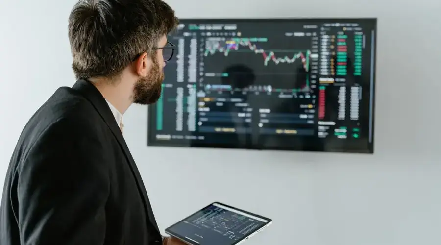A businessman examines stock market data displayed on a monitor, holding a tablet.