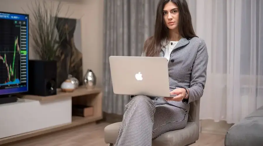 Businesswoman using laptop in cozy home office setting with stock market data on screen.