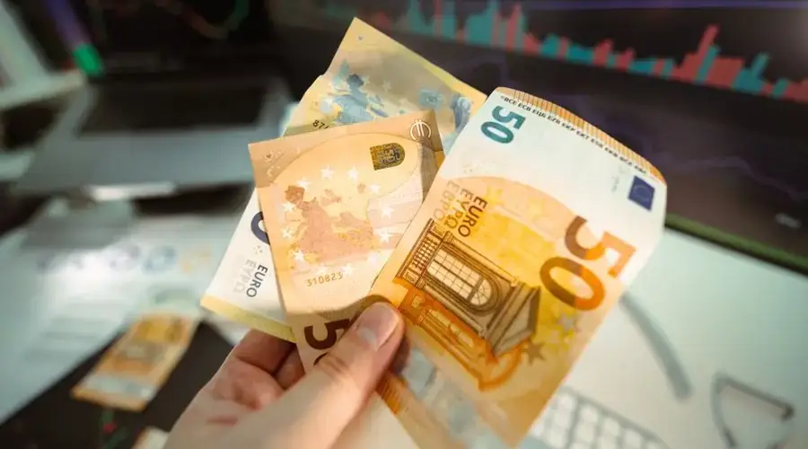 Hand holding Euro banknotes in front of a trading screen showing financial graphs.