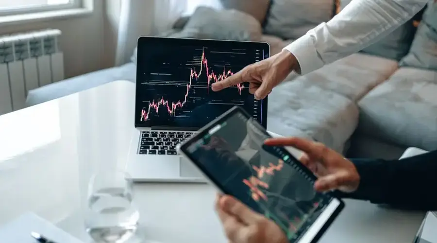 Hands analyzing stock market trends on laptop and tablet, indoors setting.