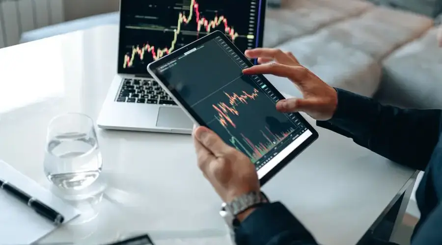 Trader analyzing stock market data on tablet with laptop in background.