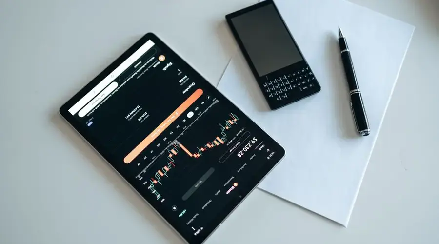 Top view of a workspace with a tablet displaying financial charts, a smartphone, pen, and paper.