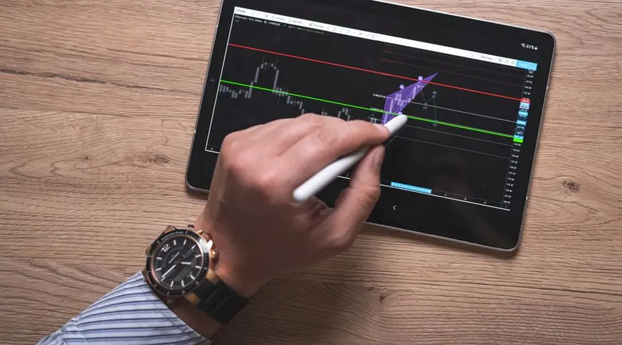 Close-up of a hand using a stylus on a digital trading app on a tablet indoors.