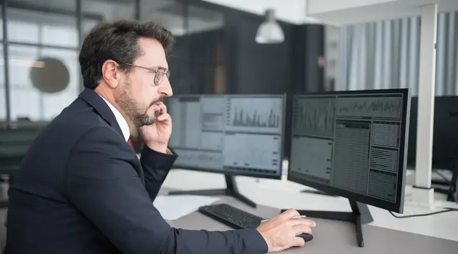 Businessman analyzing stock market data on dual monitors in a modern office setting.