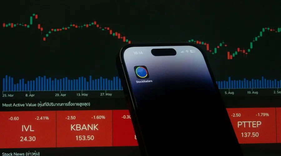 A mobile phone displaying a trading app in front of a financial market chart screen.