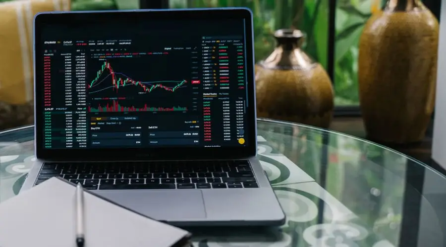 Laptop displaying cryptocurrency stocks and graphs on a glass table with a notepad.