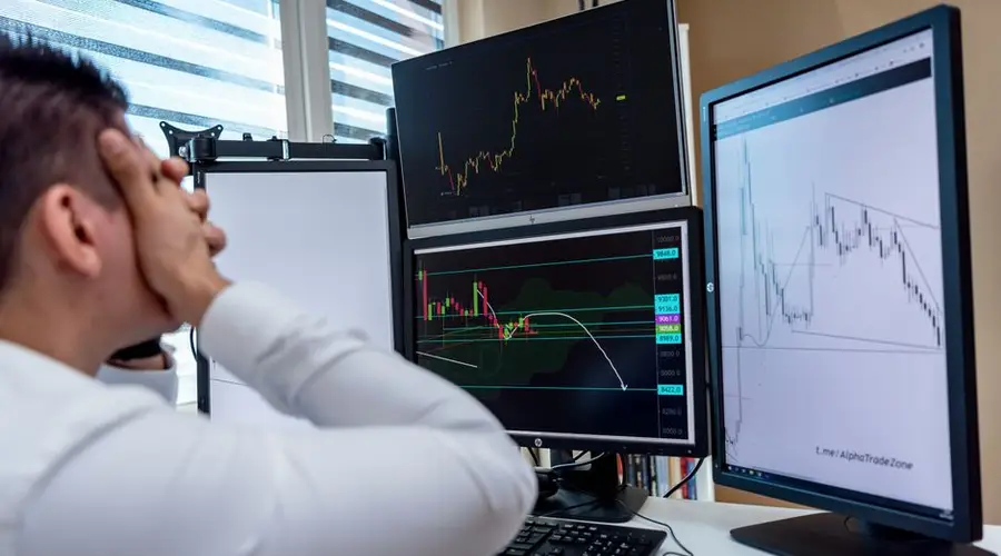 Frustrated man monitoring multiple trading graphs on computer screens in an office setting.