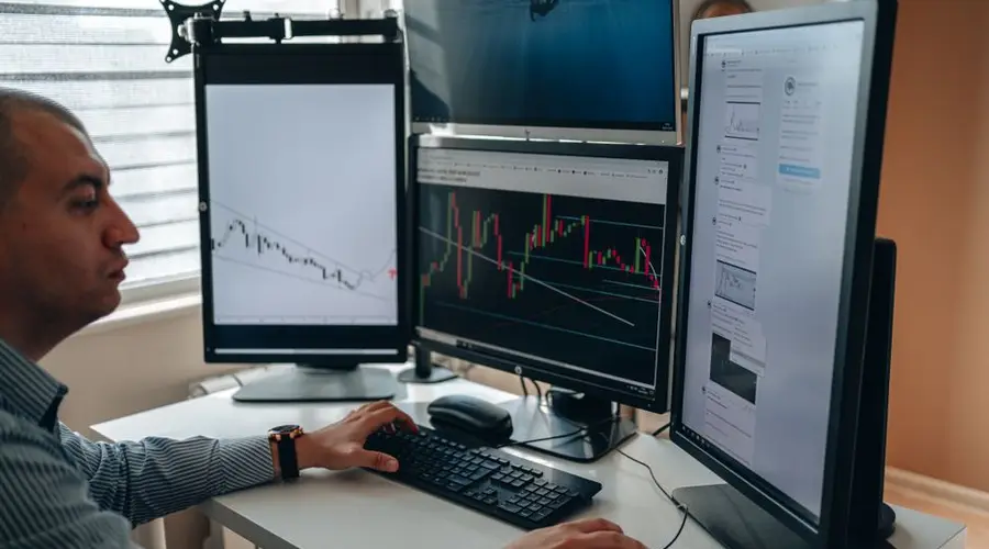 A professional in an office analyzing financial charts on multiple monitors, using advanced technology.