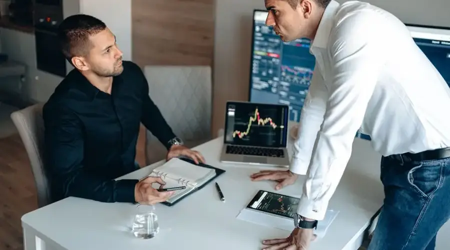 Two businessmen discuss stock market data on screens in a modern office.