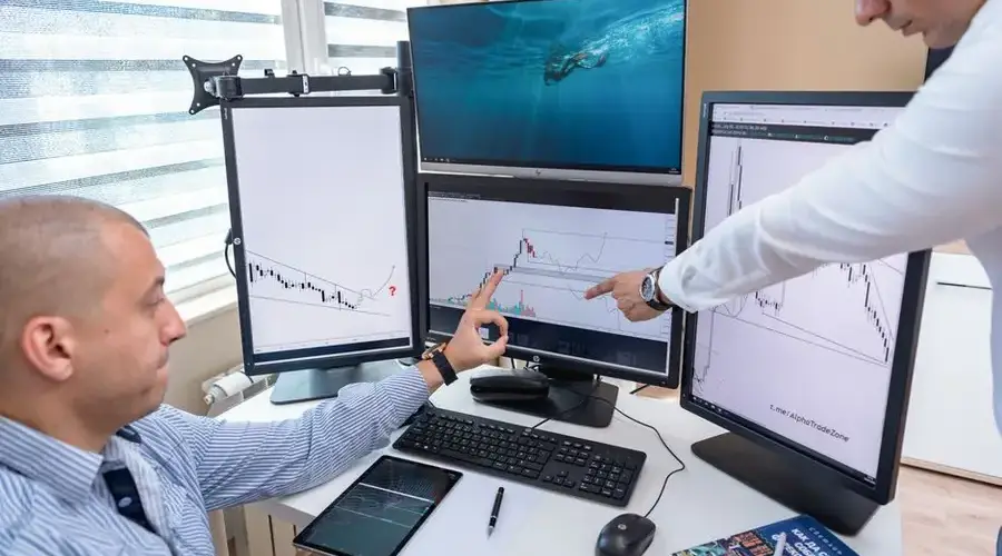 Two business professionals analyzing financial data on multiple computer screens in an office setting.