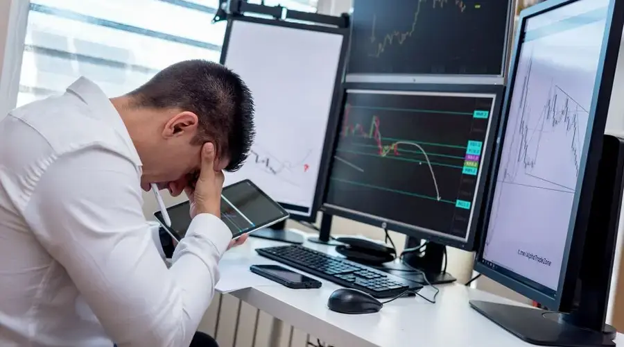 A stressed trader in an office setting analyzes market data on multiple monitors using a tablet.