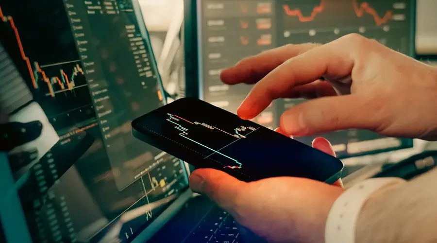 Hands interact with smartphone for analyzing cryptocurrency trading on digital screens.
