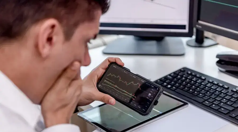 Businessman focuses on stock market analysis using smartphone and digital devices in modern workspace.
