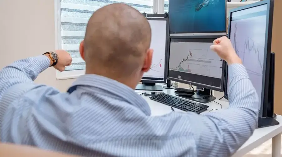 A man celebrates success at a multi-monitor workstation while analyzing stock charts.