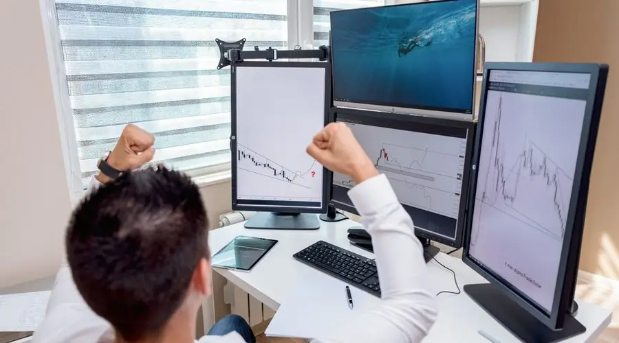 A man celebrates in front of multiple computer monitors displaying stock market graphs, indicating trading success.