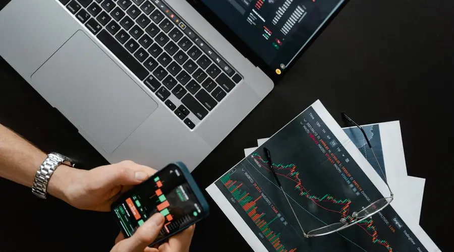 Hands using smartphone beside laptop with stock charts, showcasing digital trading.