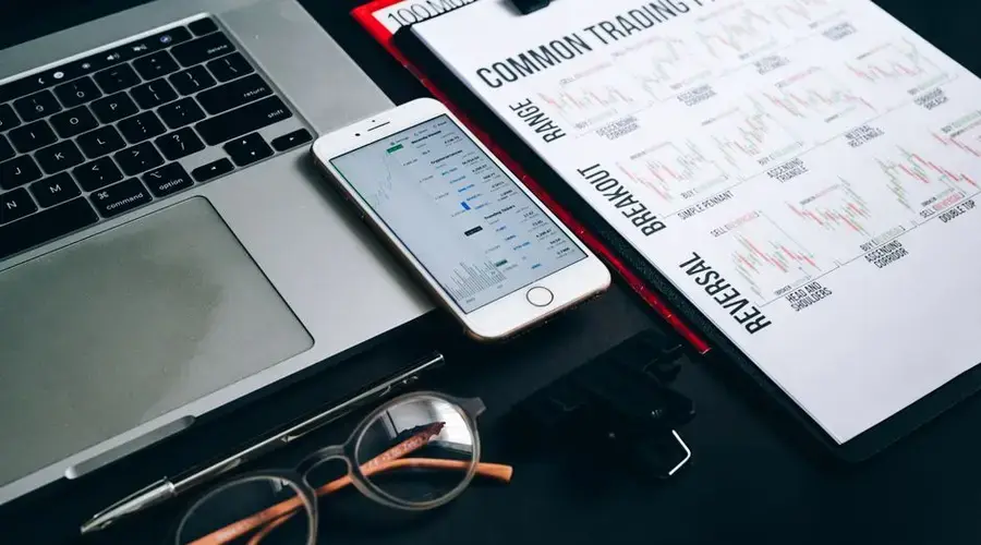 Workstation with laptop, smartphone, eyeglasses, financial charts, and pen for trading analysis.