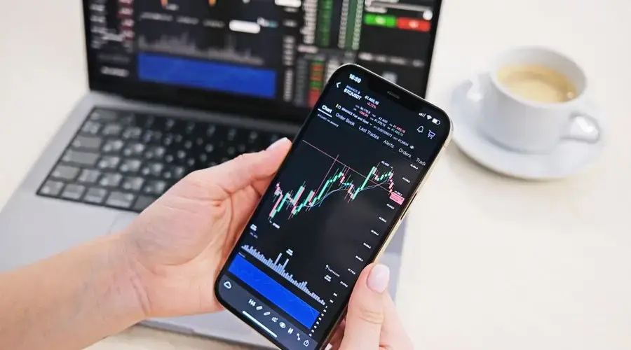 Close-up of hands holding smartphone displaying stock charts with laptop in the background on a desk.