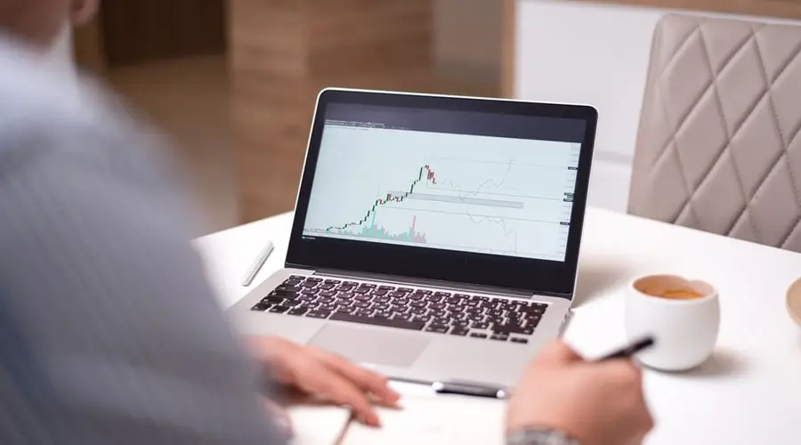 A person in a home office analyzing stock market data on a laptop with a coffee cup beside them.