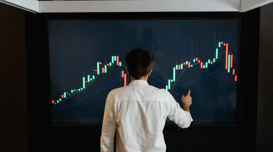 Adult male reviewing stock market data on a large display screen indoors.