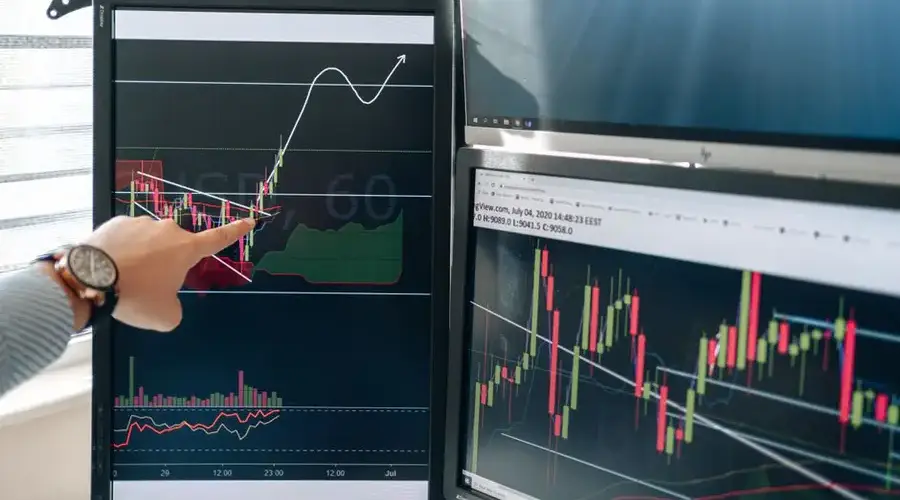 Person pointing at stock market graphs on dual monitors in a modern workspace.