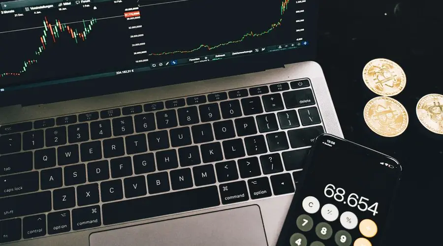 A laptop with trading charts, smartphone calculator, and bitcoin coins depicting cryptocurrency trading.