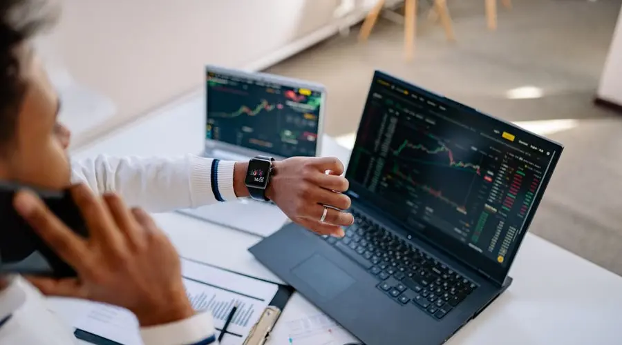 Man multitasking with a smartwatch and laptop analyzing stock market charts at the office.